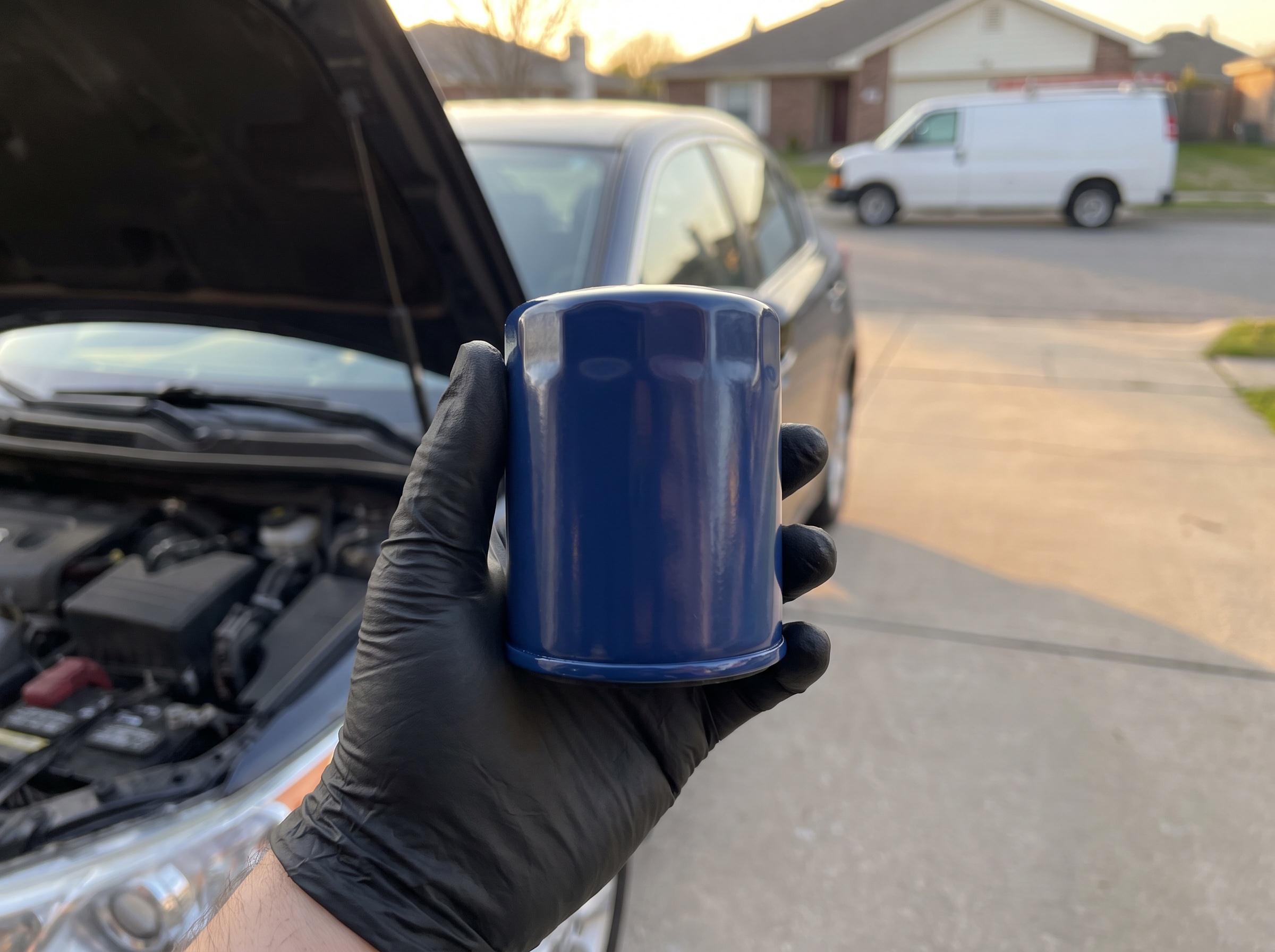 A gloved hand holding a new oil filter beside an open hood in a driveway
